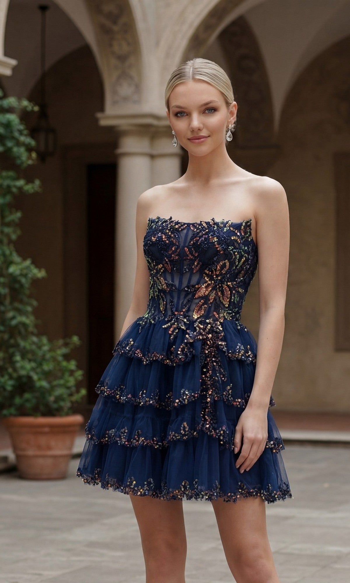 A woman stands outdoors in a navy blue, strapless Nox Anabel G7043 short homecoming dress with layered tulle and sequin embellishments. The scene includes arched architecture and potted plants in the background.
